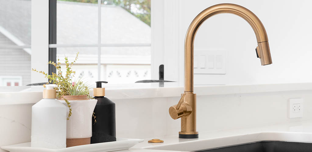 A beautiful sink in a remodeled modern farmhouse kitchen with a gold faucet, black apron or farmhouse sink, white granite, and a tiled backsplash.